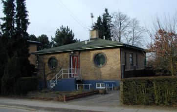Eksempel på en klassisk bungalow i gule sten med pyramidetag, grønt tagpap, central skorsten og en trappe i beton, der fører op til hoveddøren.