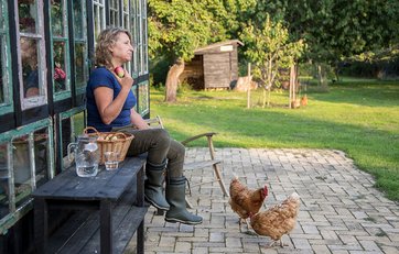 Birgitte Svennevig sidder på bænk opad sit hus og spiser æble, mens høns går omkring på terassen